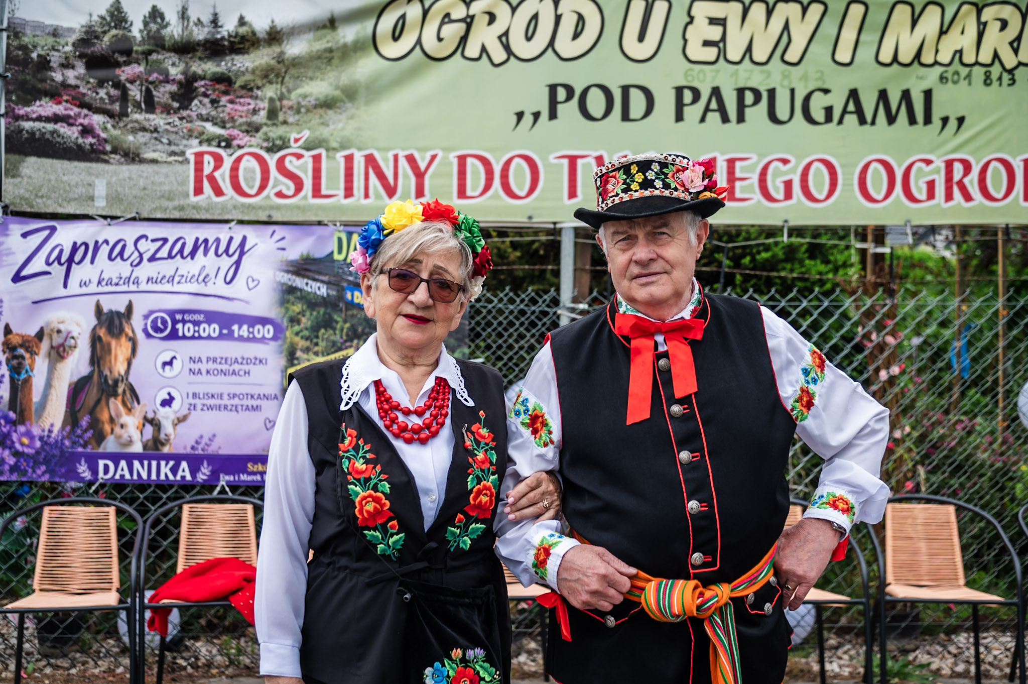 Uczestnicy wydarzenia w tradycyjnych strojach ludowych na tle banerów Szkółki Ogrodniczej Danika