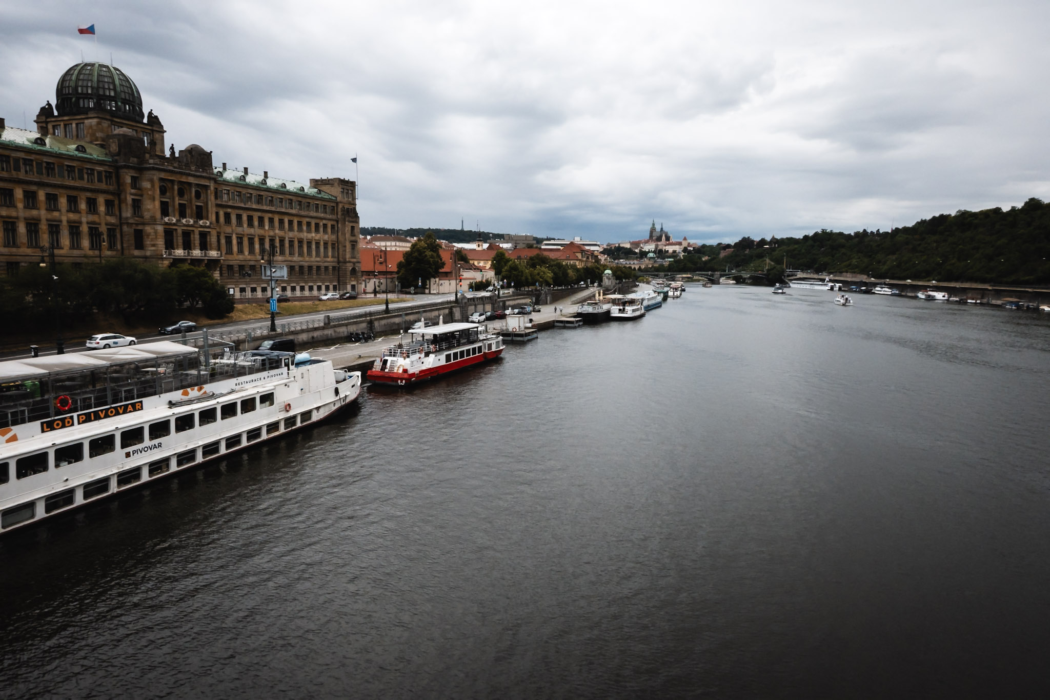 Panorama Pragi z widokiem na Wełtawę, mosty, statki i zabudowę miasta pod pochmurnym niebem